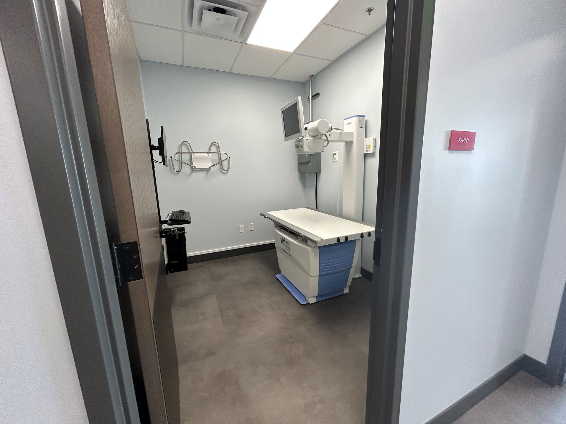 Exam room with X-ray machine, table, and medical equipment. Gray walls, dark floor, doorway view.