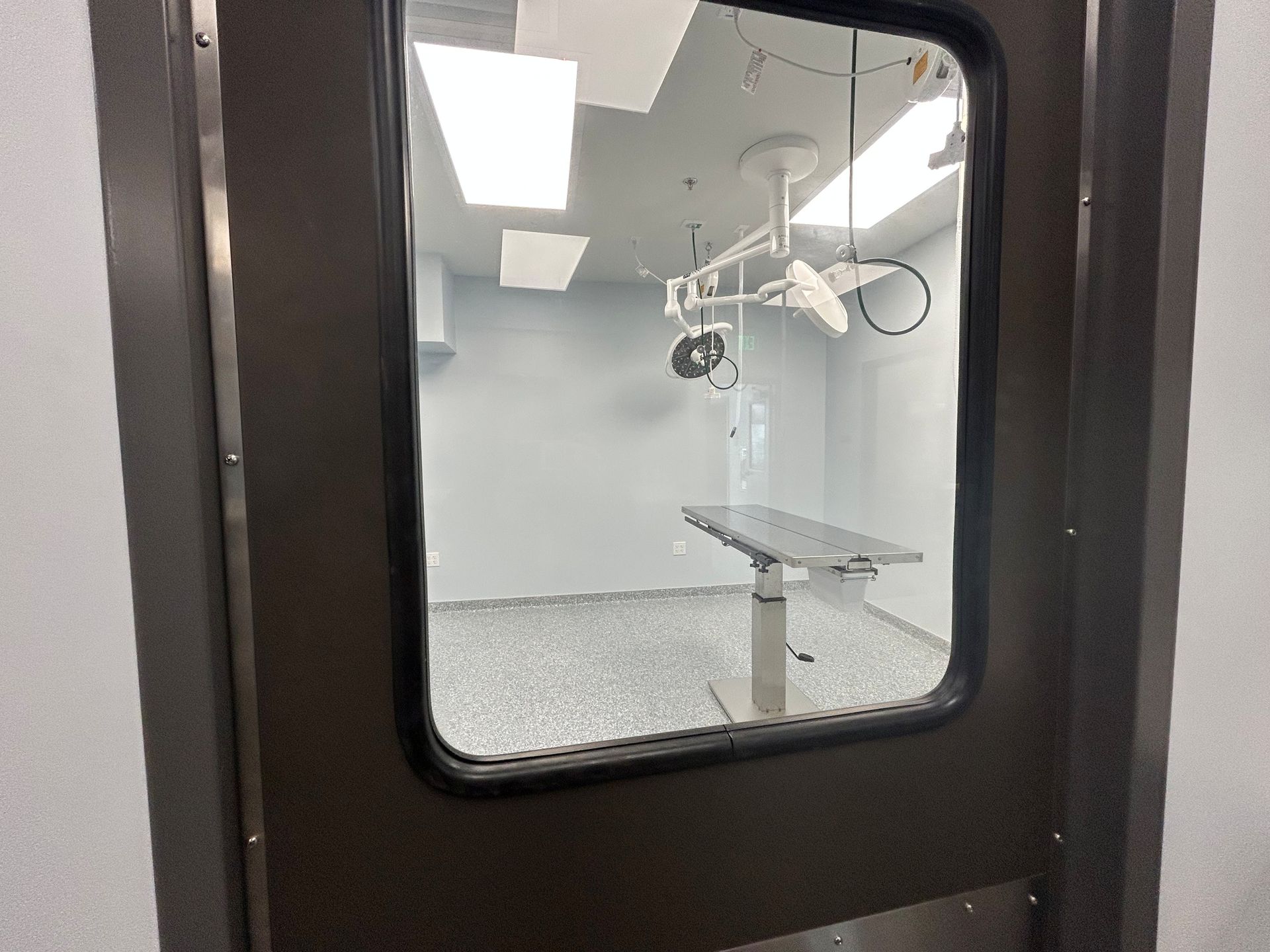 A medical operating room seen through a door window. A surgical table and overhead lights are visible inside.