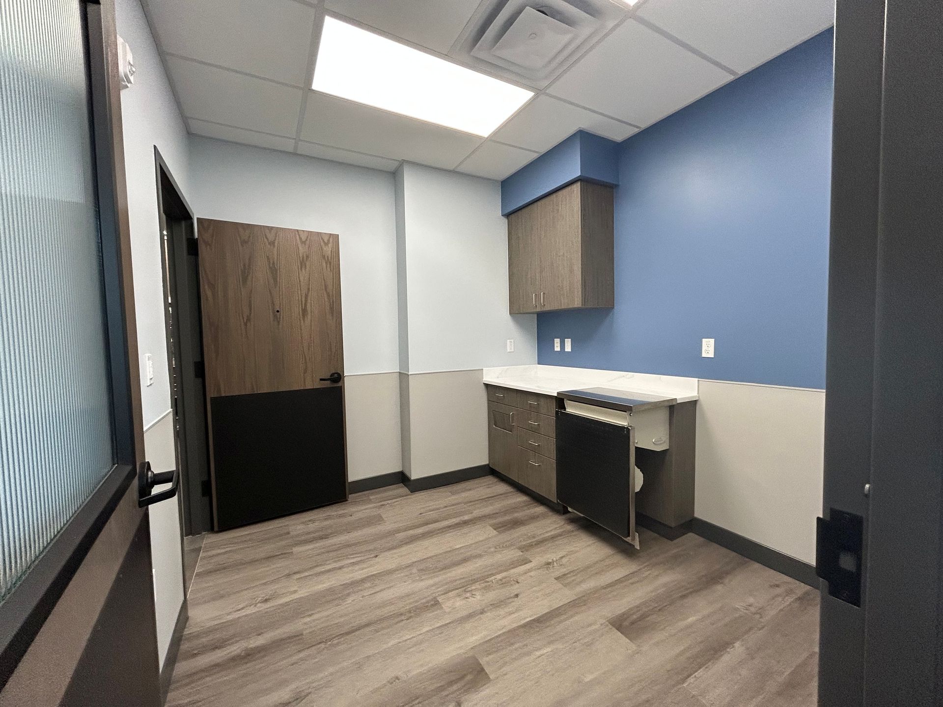 Empty office room with blue and tan walls, cabinets, sink, and wood-look flooring.