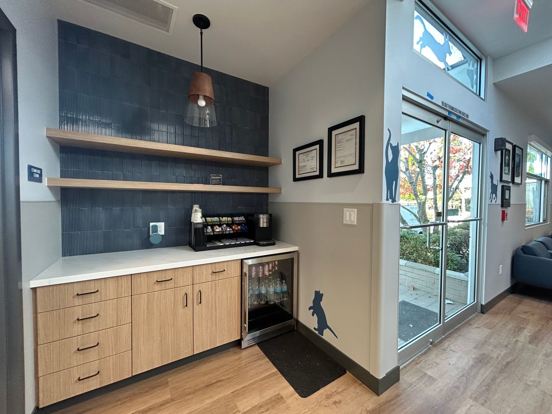 Coffee station in a waiting area with wooden cabinetry, shelves, and a refrigerator; a glass door leads outside.