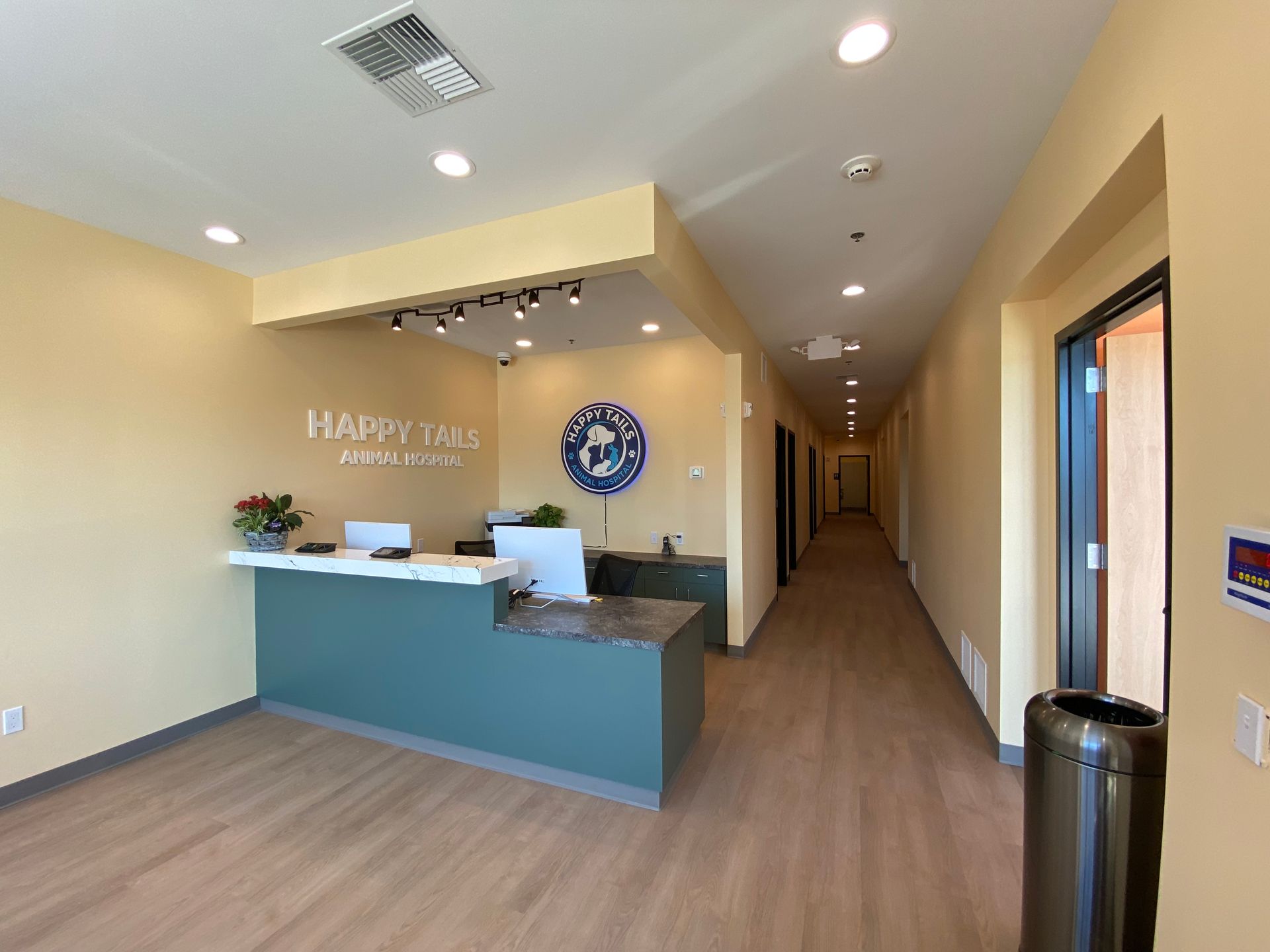 Reception area with teal front desk and long hallway. Happy Tails sign, wooden floors, beige walls.