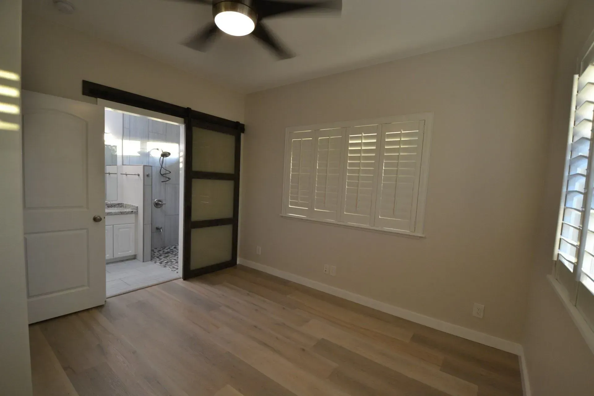 Empty bedroom with light wood floors, sliding door to bathroom, window with shutters.