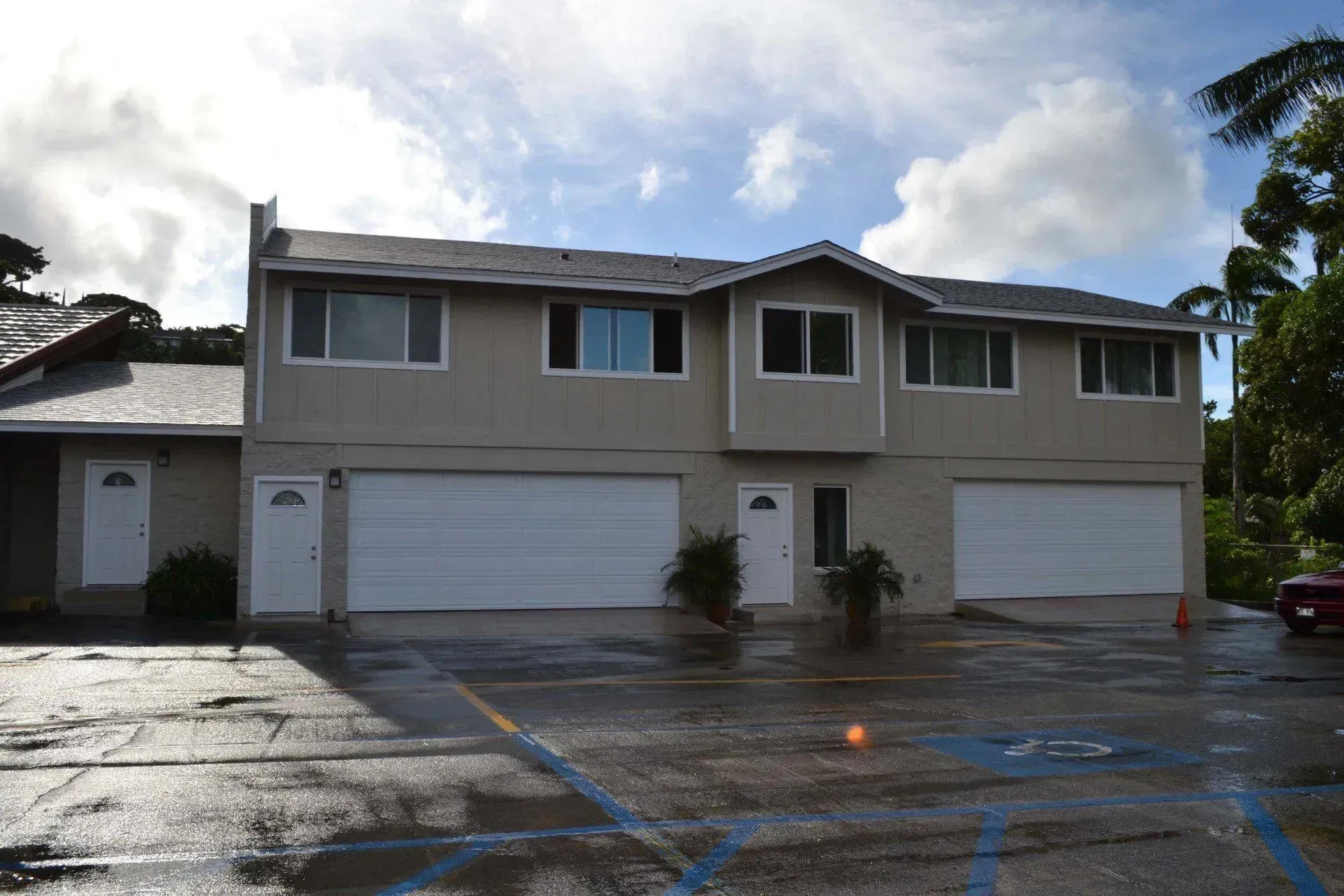 Two-story beige building with garage doors, front door, and windows. Overcast sky, wet parking lot.