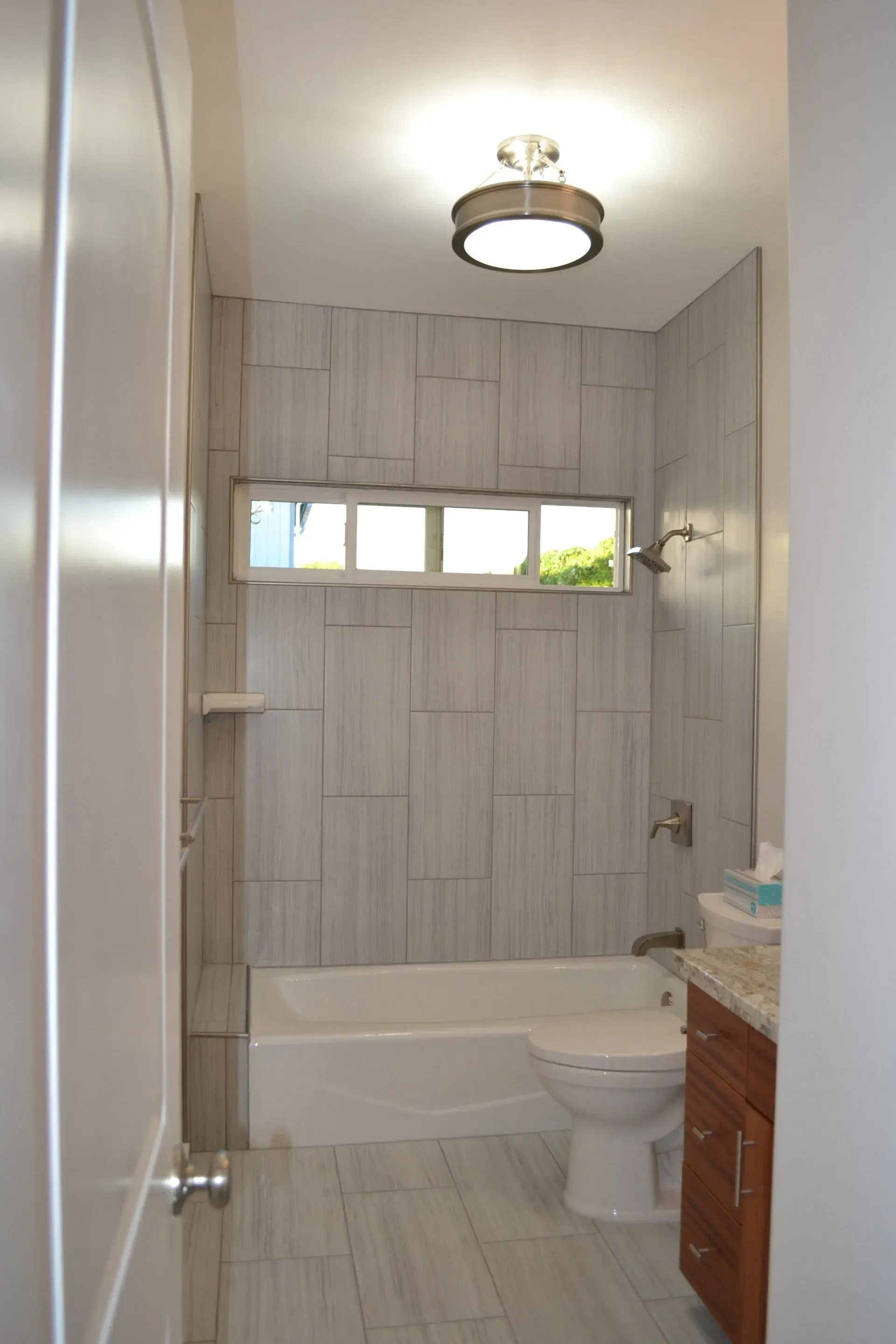 Bathroom with light wood-look tile, a white bathtub, toilet, and sink, with a window and circular ceiling light.