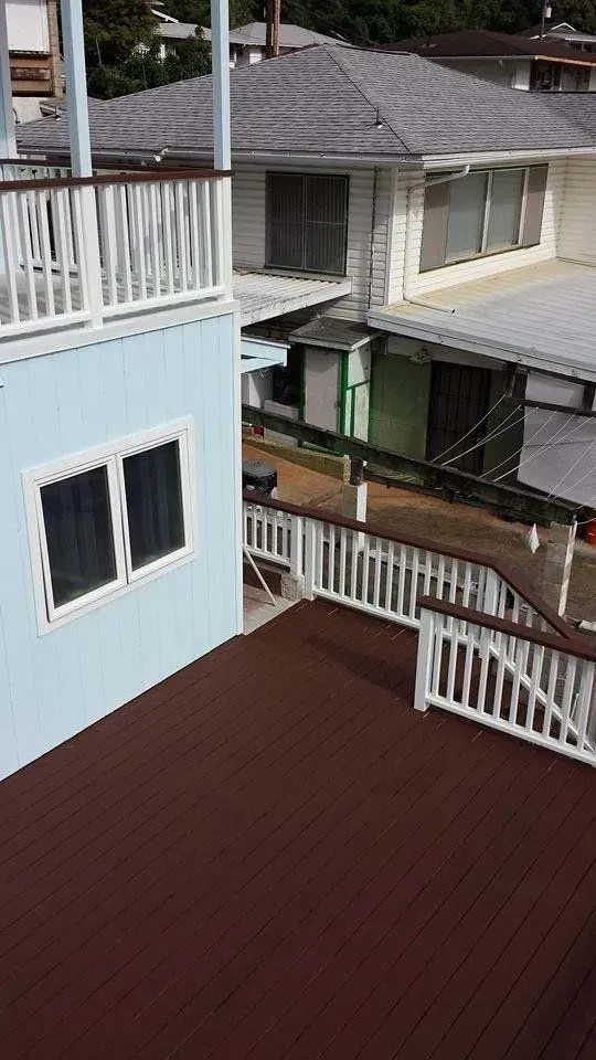 Brown wooden deck with white railing, light blue building, and houses in the background.