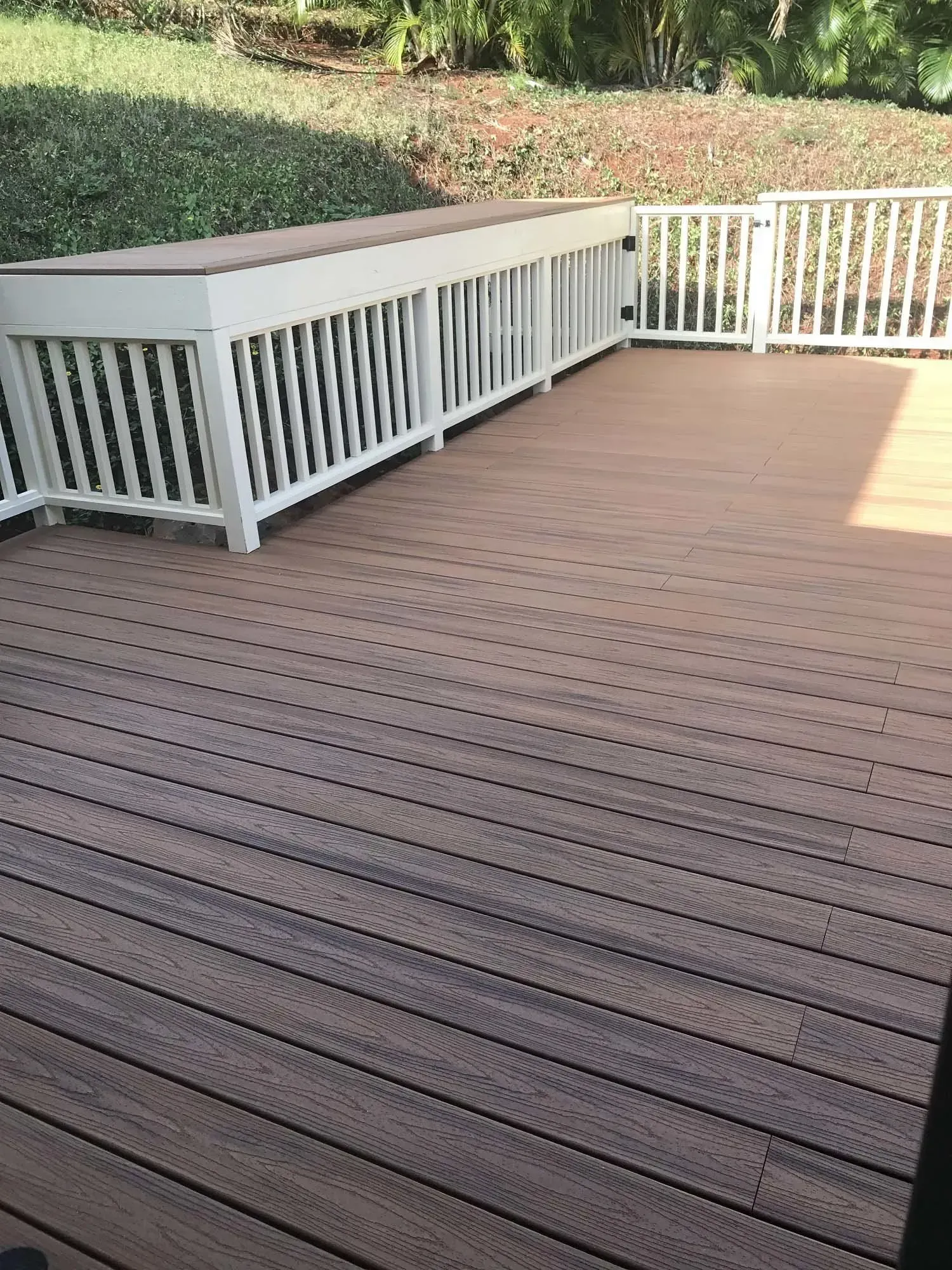A brown composite deck with white railings and a built-in bench, with a green hillside in the background.