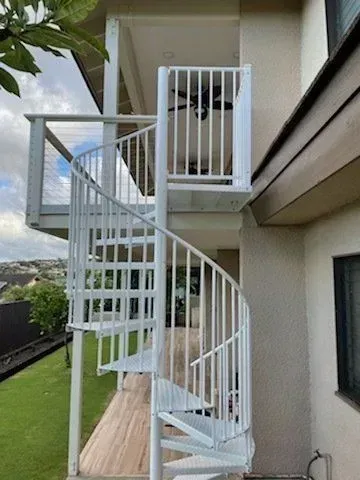 White spiral staircase leads up to a balcony, attached to a house with a grass yard.