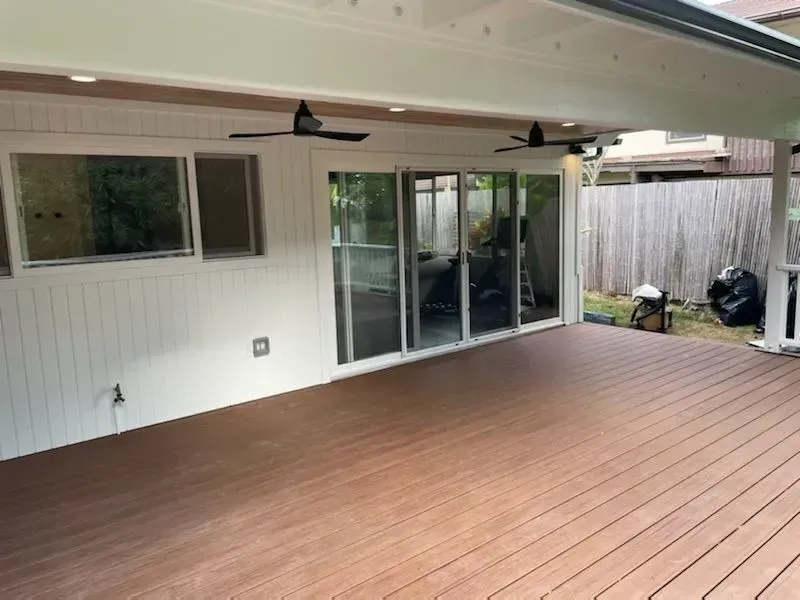 Covered patio with brown decking, sliding glass doors, white walls, and ceiling fans.
