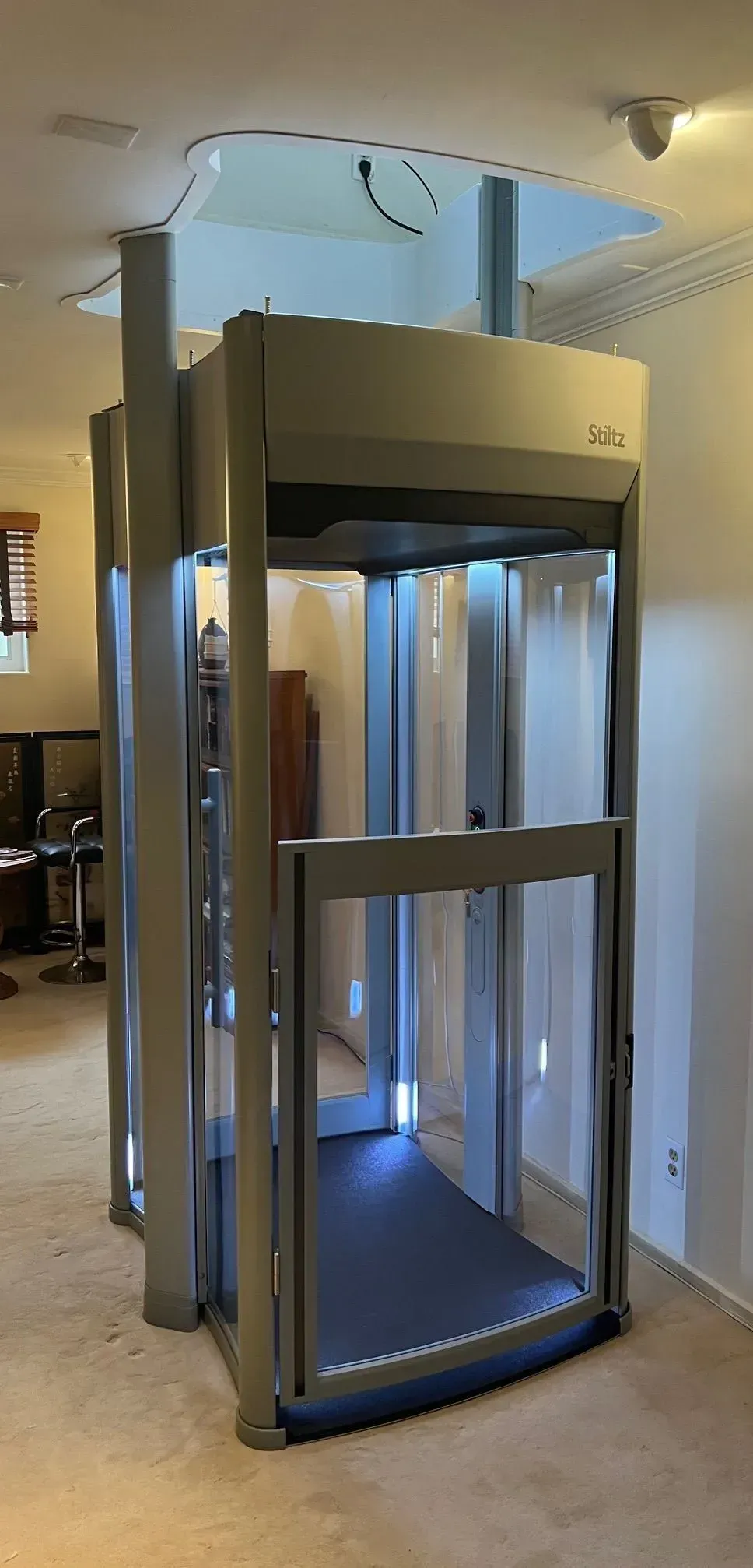 Indoor residential elevator, metal frame with glass panels. Elevator platform has blue lighting. Beige carpeted floor and walls.