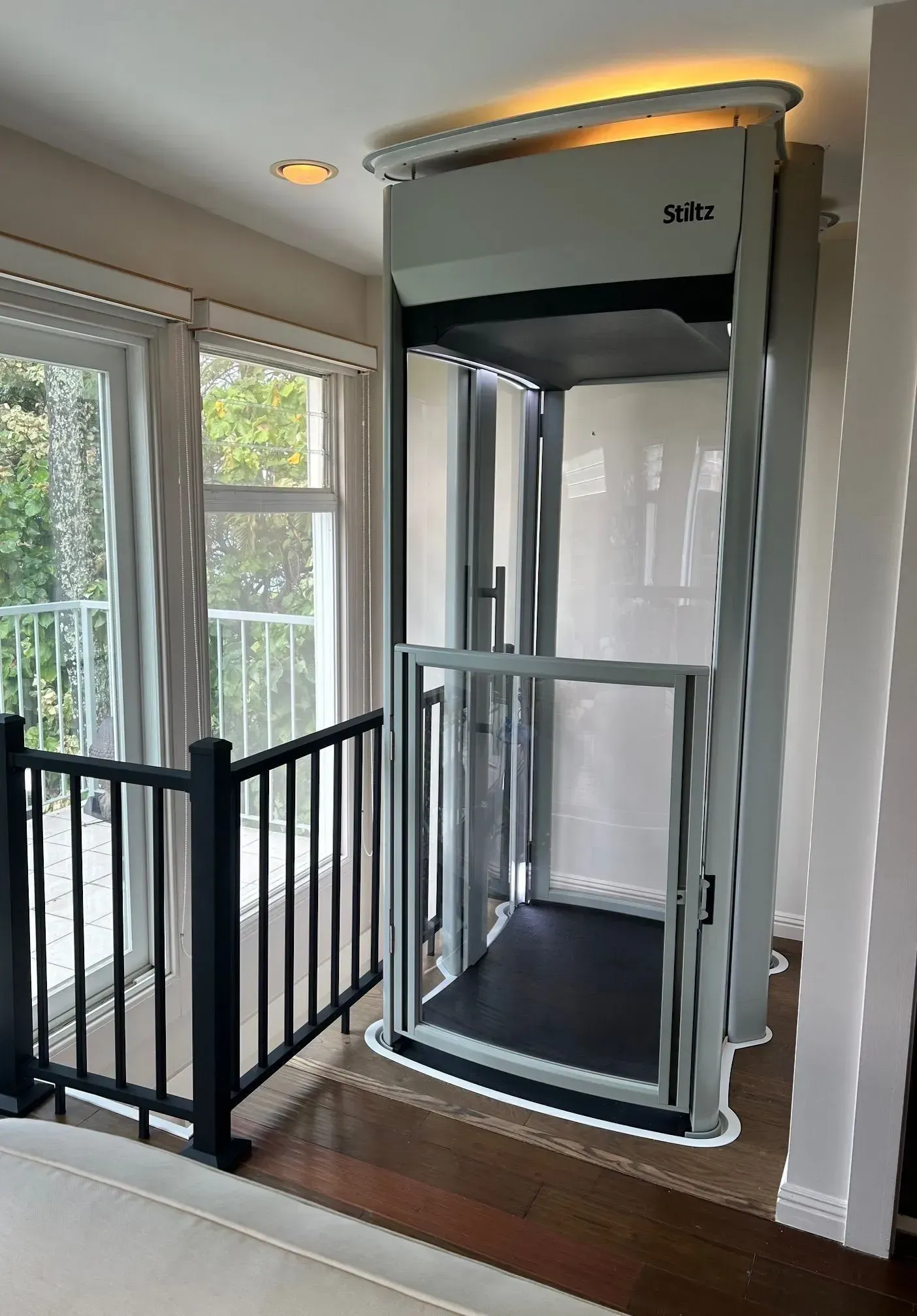 Gray home elevator installed beside a balcony. The elevator has a glass door, and a black floor.