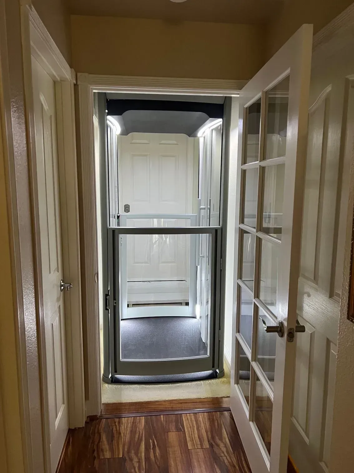 Hallway with a home elevator; white doors and trim, wood floor, glass enclosure.