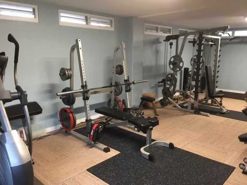 Home gym with exercise equipment: weights, bench, and elliptical. Light blue walls, tan flooring.