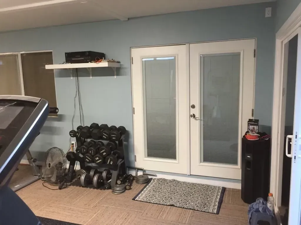 Home gym with dumbbells, treadmill, and double doors. Light blue walls and beige carpet.