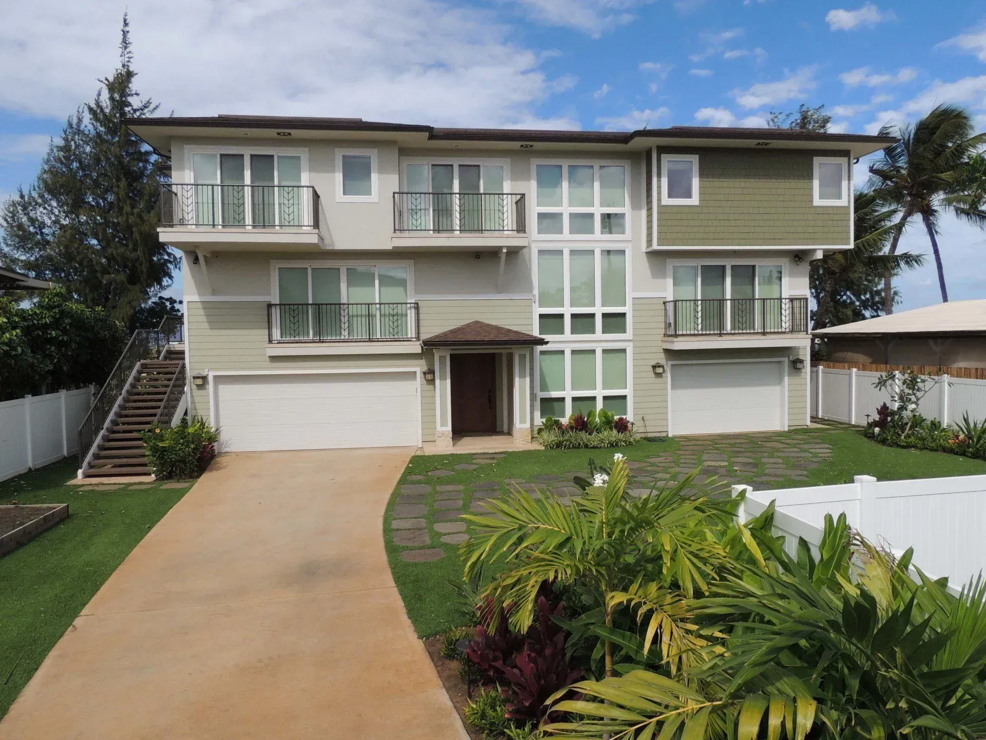Two-story beige house with balconies, large windows, and a driveway. Palm trees and grass in front.