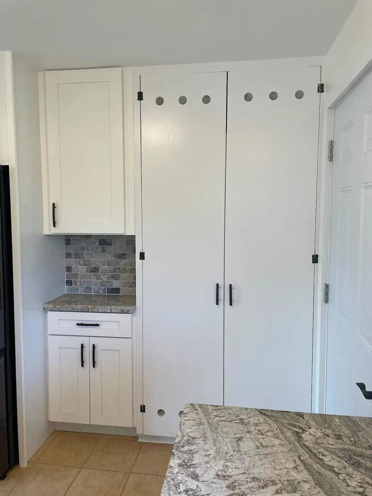 White kitchen cabinets with black hardware and a gray countertop, next to a white door.