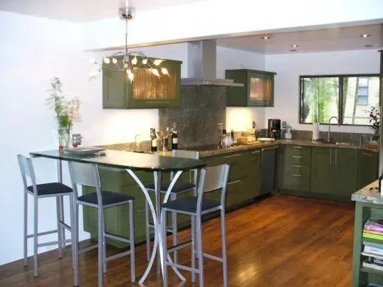 Modern kitchen with olive-green cabinets, hardwood floors, and a glass-topped bar with metal stools.