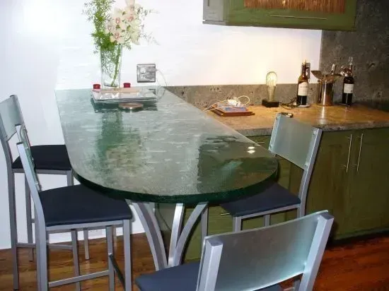 Green glass-topped bar table with three bar stools in a kitchen. Flowers and wine bottles are on the countertop.