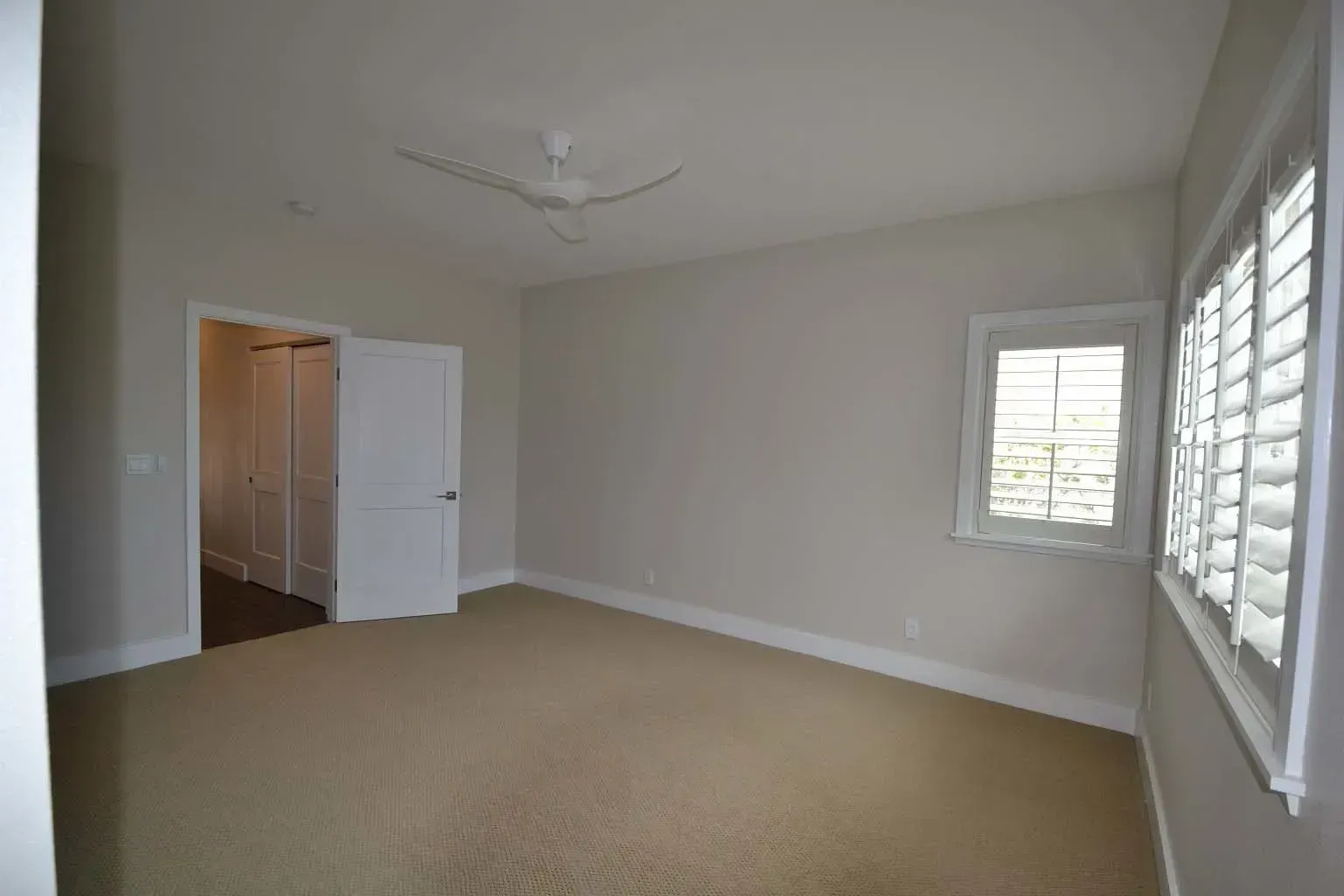 Empty bedroom with tan carpet, white walls, closet door, and shuttered window.