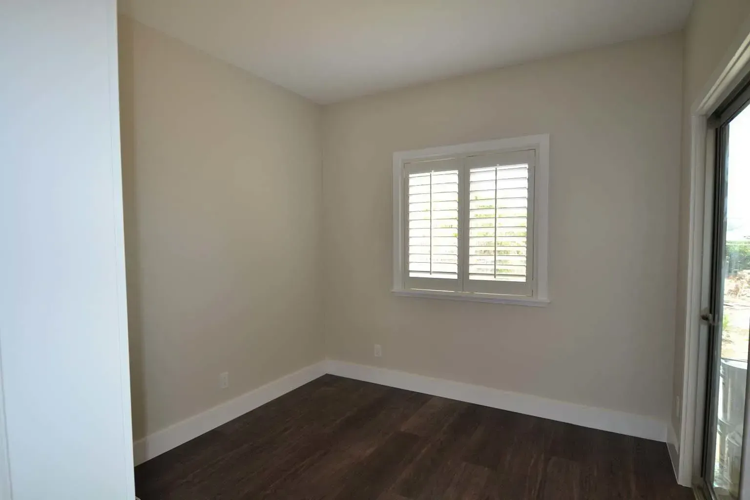 Empty room with dark wood floor, light beige walls, and window with shutters.