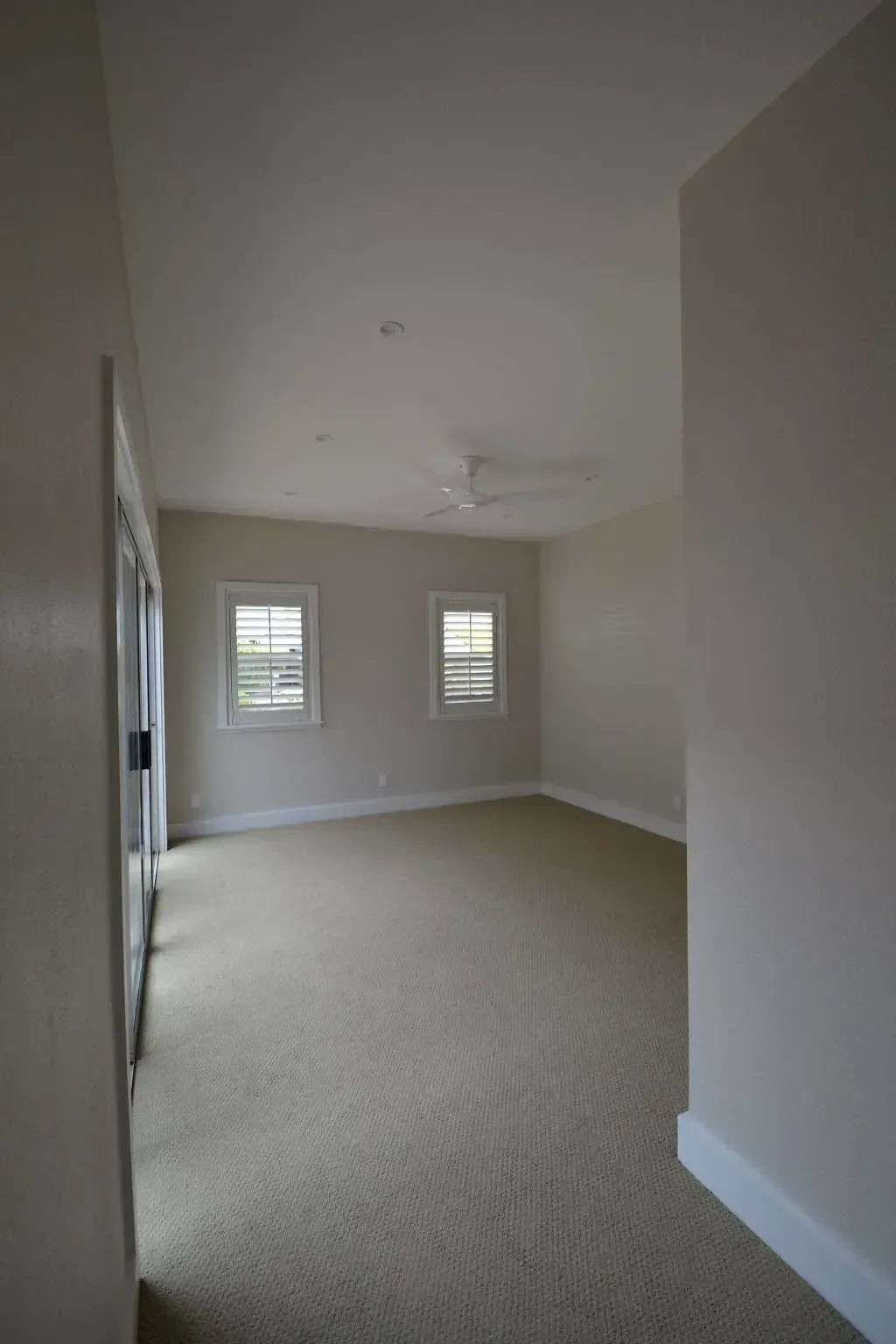 Empty bedroom with beige walls, carpet, two windows, and a ceiling fan.