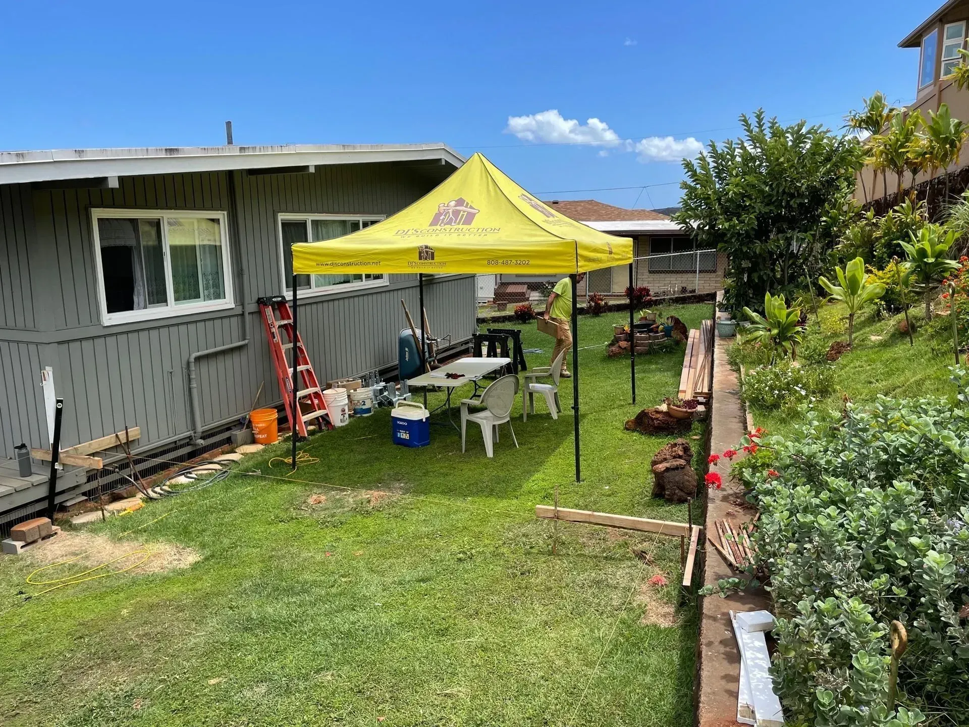 A backyard with a yellow tent set up. Tables, chairs, and tools are visible.