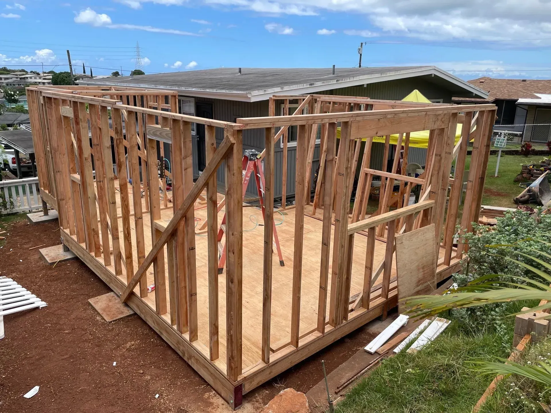 Wooden frame of a building under construction, exterior view, with sky and houses in background.