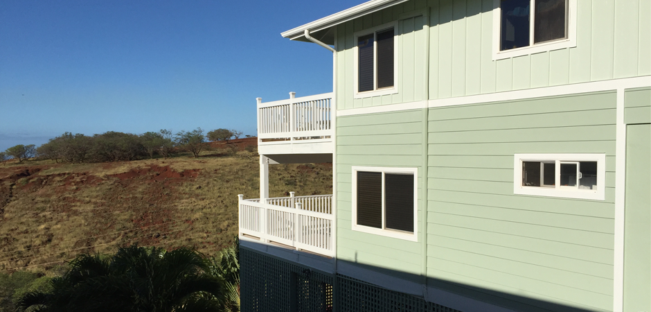Two-story light green house with white railing and windows, set against a hill and blue sky.