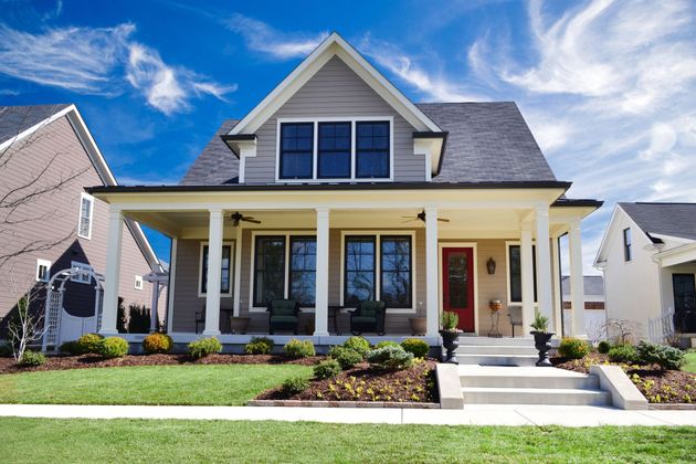 A two-story suburban house with a large front porch, beige siding, gray roof, and a red front door under a sunny blue sky.
