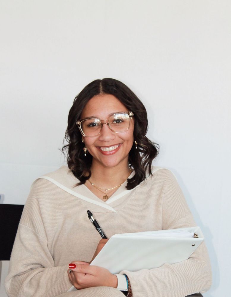 Nikki Bates smiling, writing on a clipboard, wearing glasses and a beige sweater, against a white background.