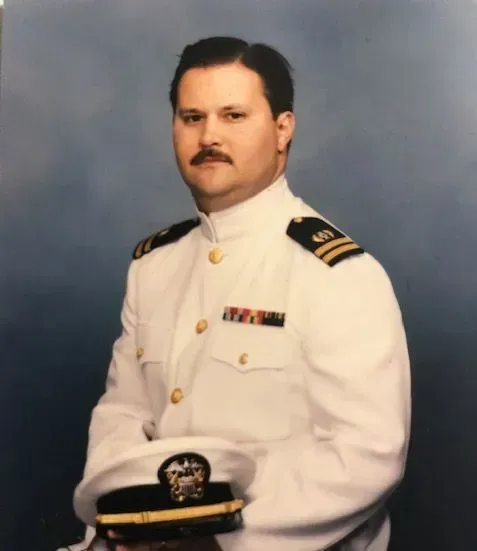 Man in white naval uniform, holding hat, smiling, against a blue background.