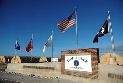 Camp Justice sign with flags, including U.S. flag, and tan tent structures in the background.