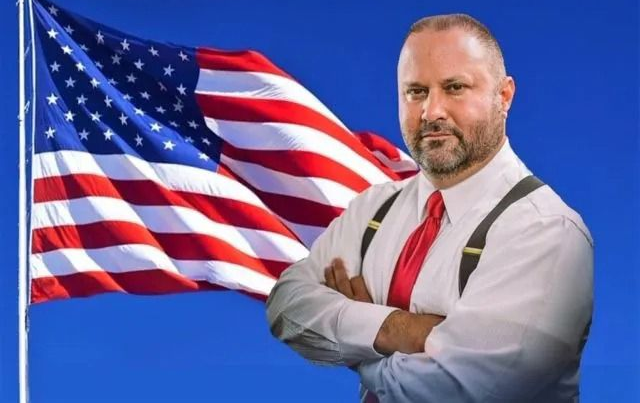 Man with arms crossed, in front of American flag, wearing white shirt, red tie, and suspenders.
