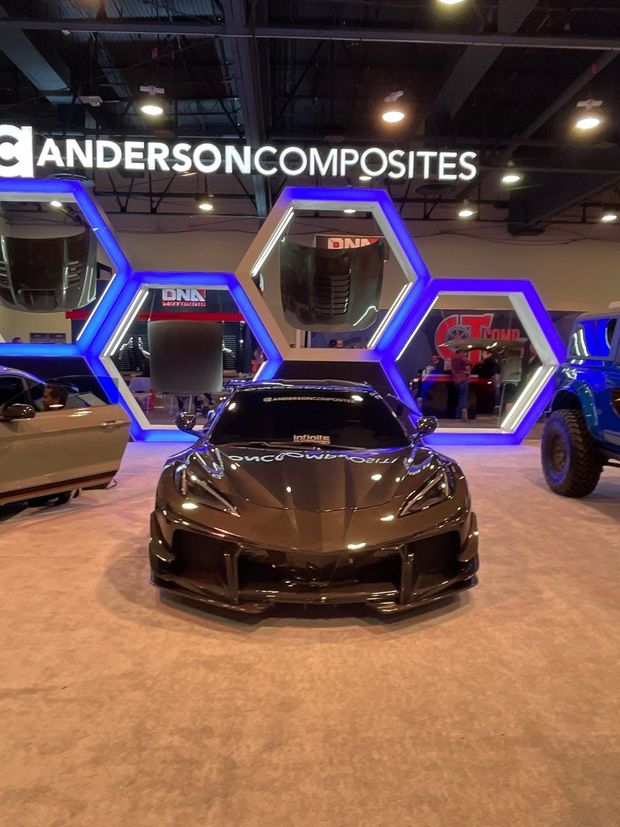 A dark-colored sports car parked at an Anderson Composites booth under illuminated hexagonal displays at a trade show.