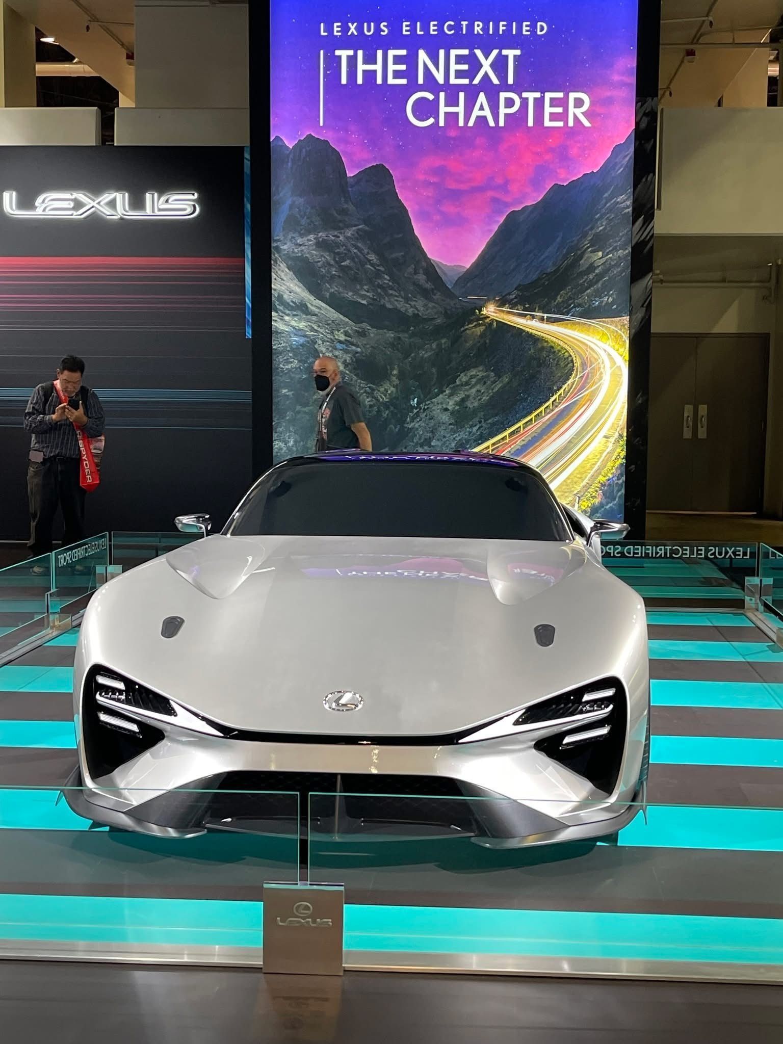 A white Lexus concept sports car on a blue-striped display floor in front of a digital screen reading "The Next Chapter."