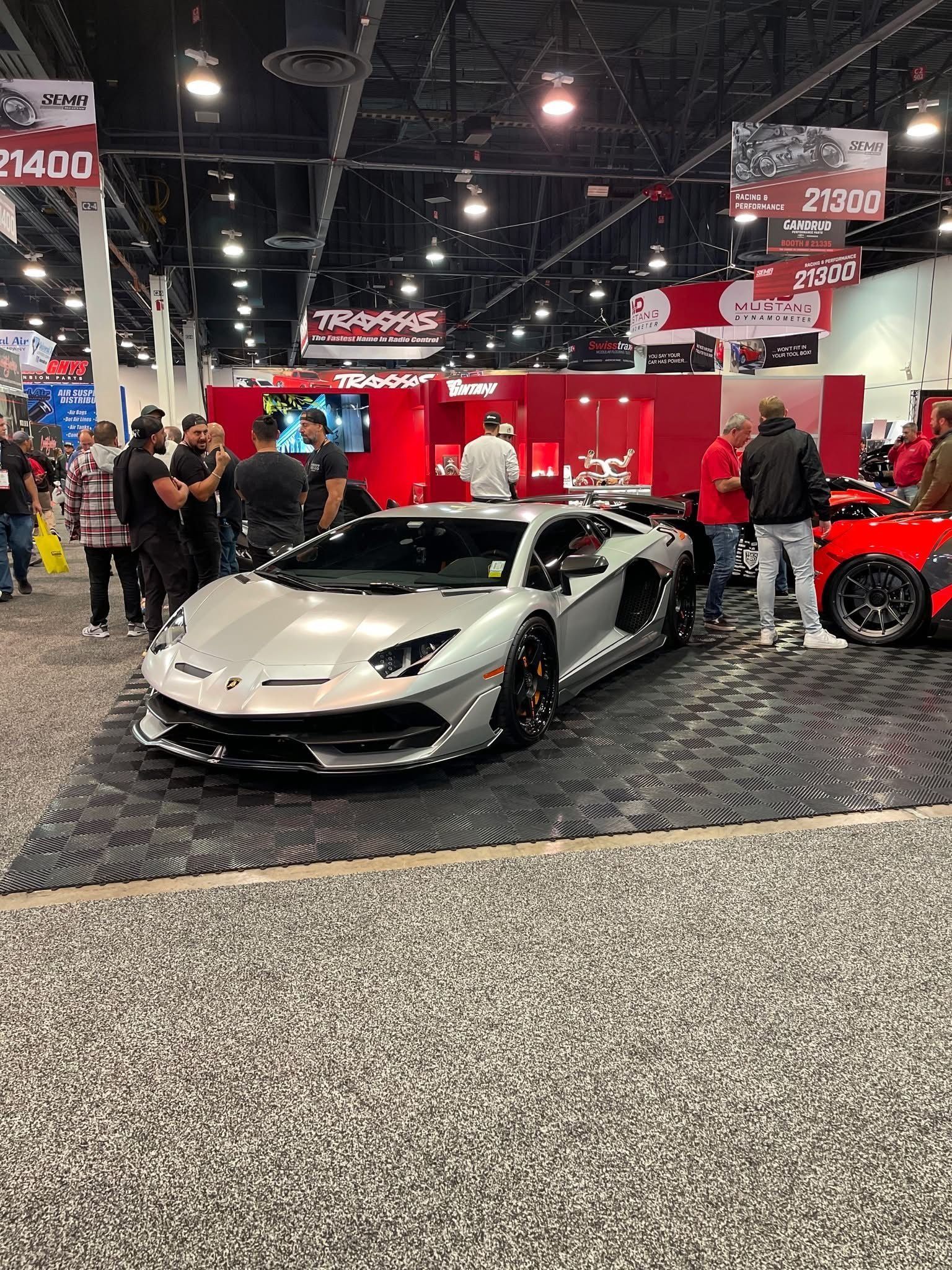 A silver Lamborghini Aventador displayed on a textured floor at a trade show exhibit with people standing nearby.