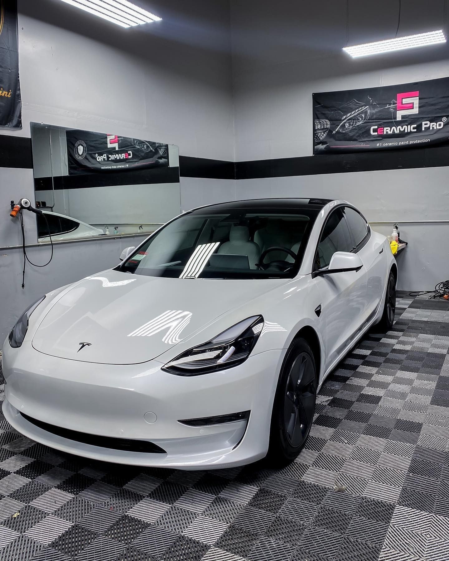 A white Tesla Model 3 parked in a professional detailing studio with checkered flooring and signage on the wall.