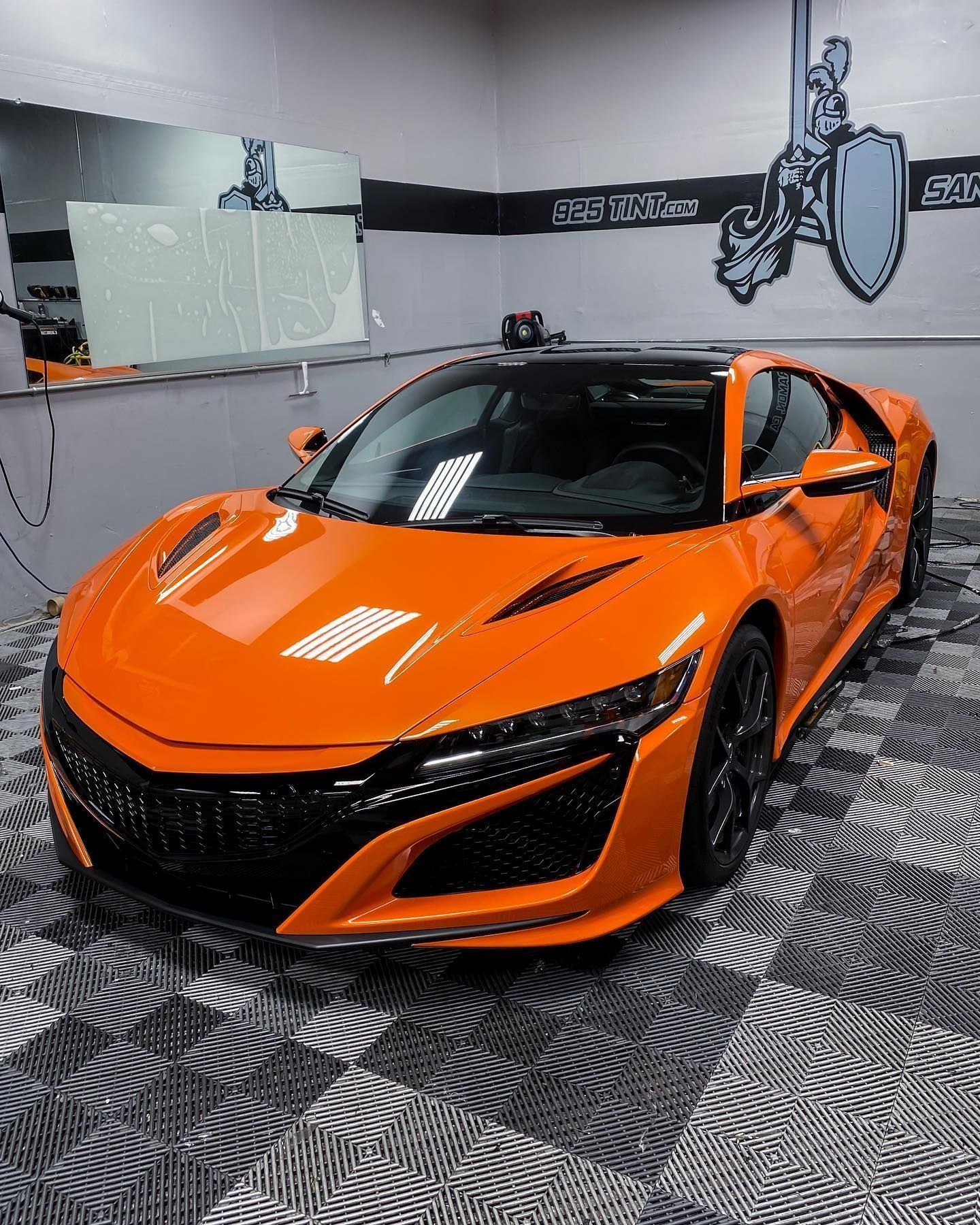 A bright orange Acura NSX sports car parked inside a professional detailing garage with a checkered floor.