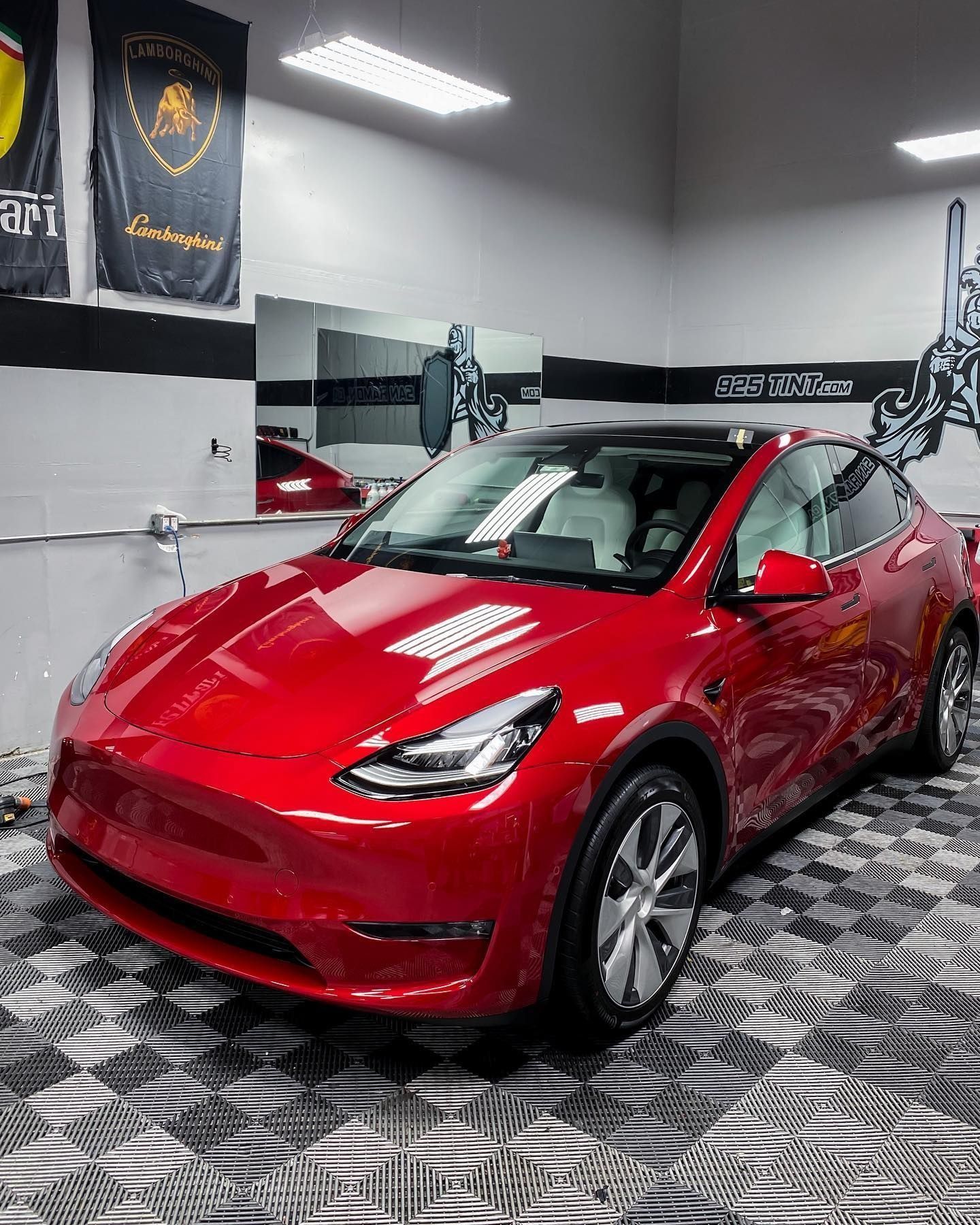A red Tesla Model Y parked on a checkered floor in a well-lit garage with banners on the wall.