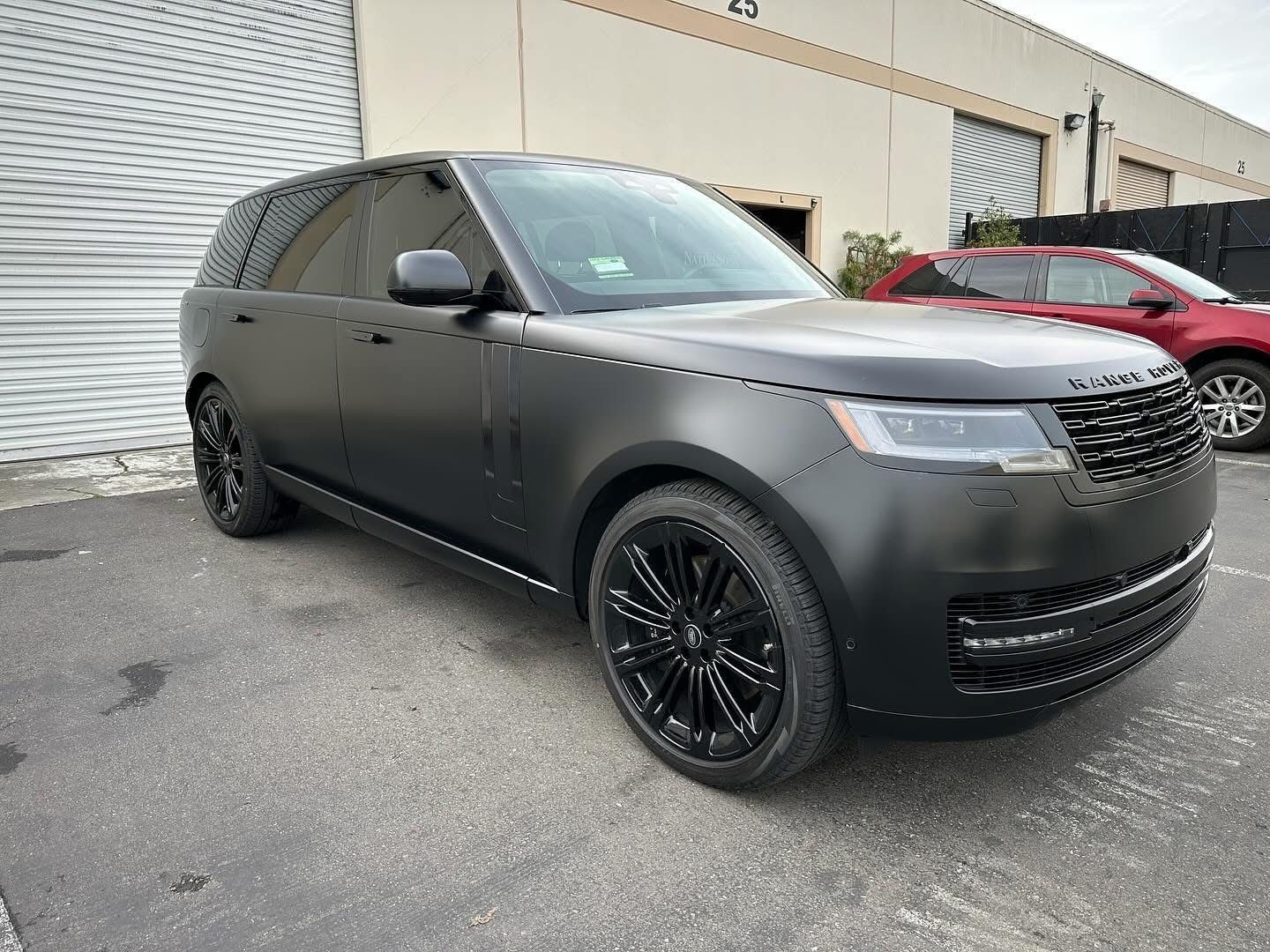 A matte black Range Rover parked on asphalt in front of a building, with a partial view of a red car in the background.