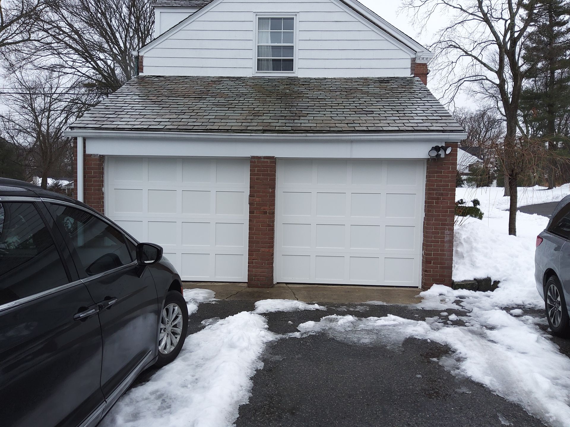 Snow-covered garage with white doors, flanked by parked cars and a cleared driveway
