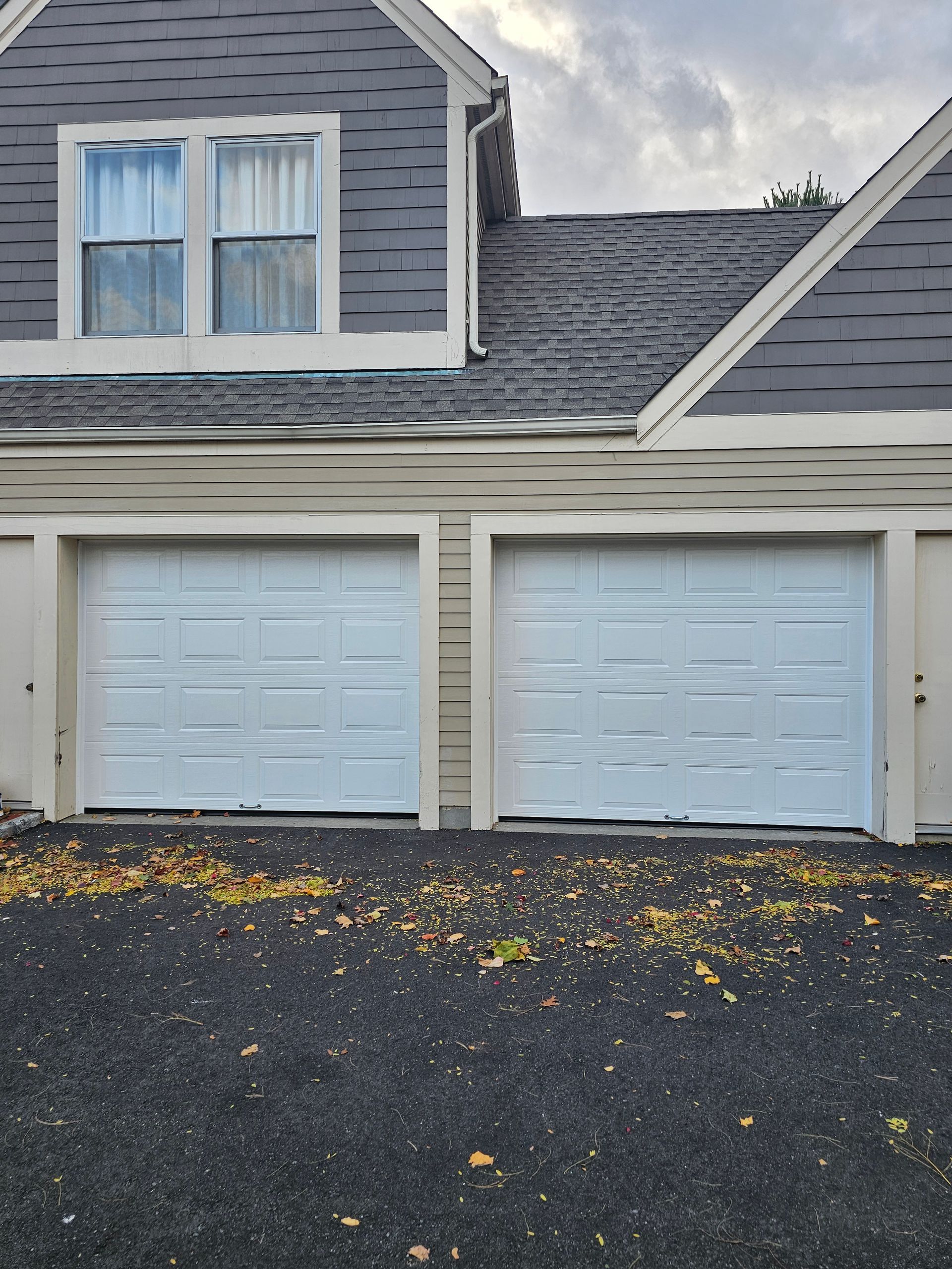 Two white garage doors on a gray house with dark shingle roof and black driveway with scattered leaves