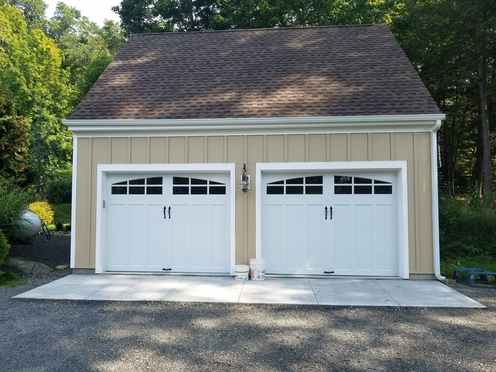 Two white garage doors on a beige detached garage with a shingled roof and driveway