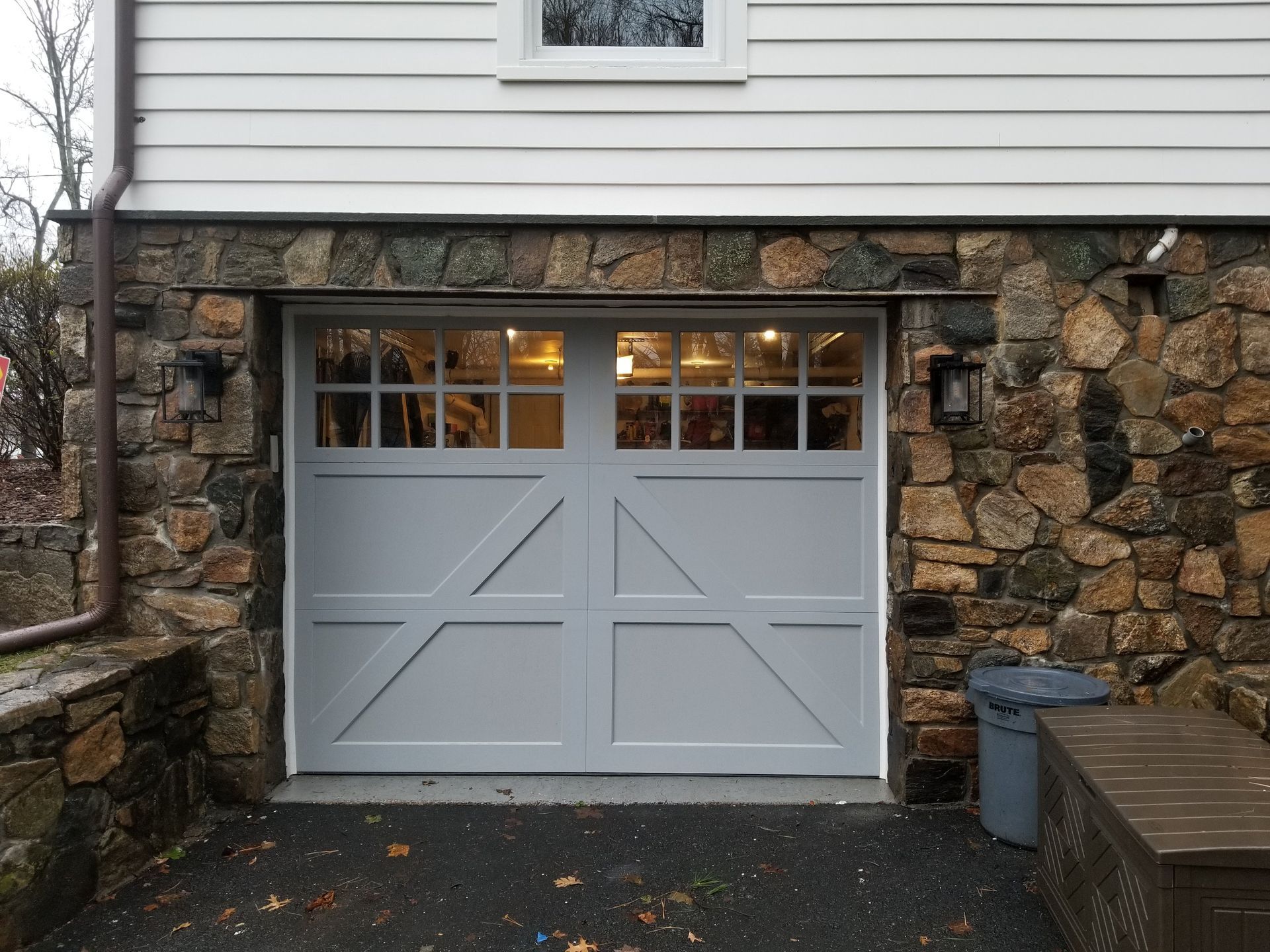 White garage door set in a stone wall beneath a house siding, with a small windowed top section.