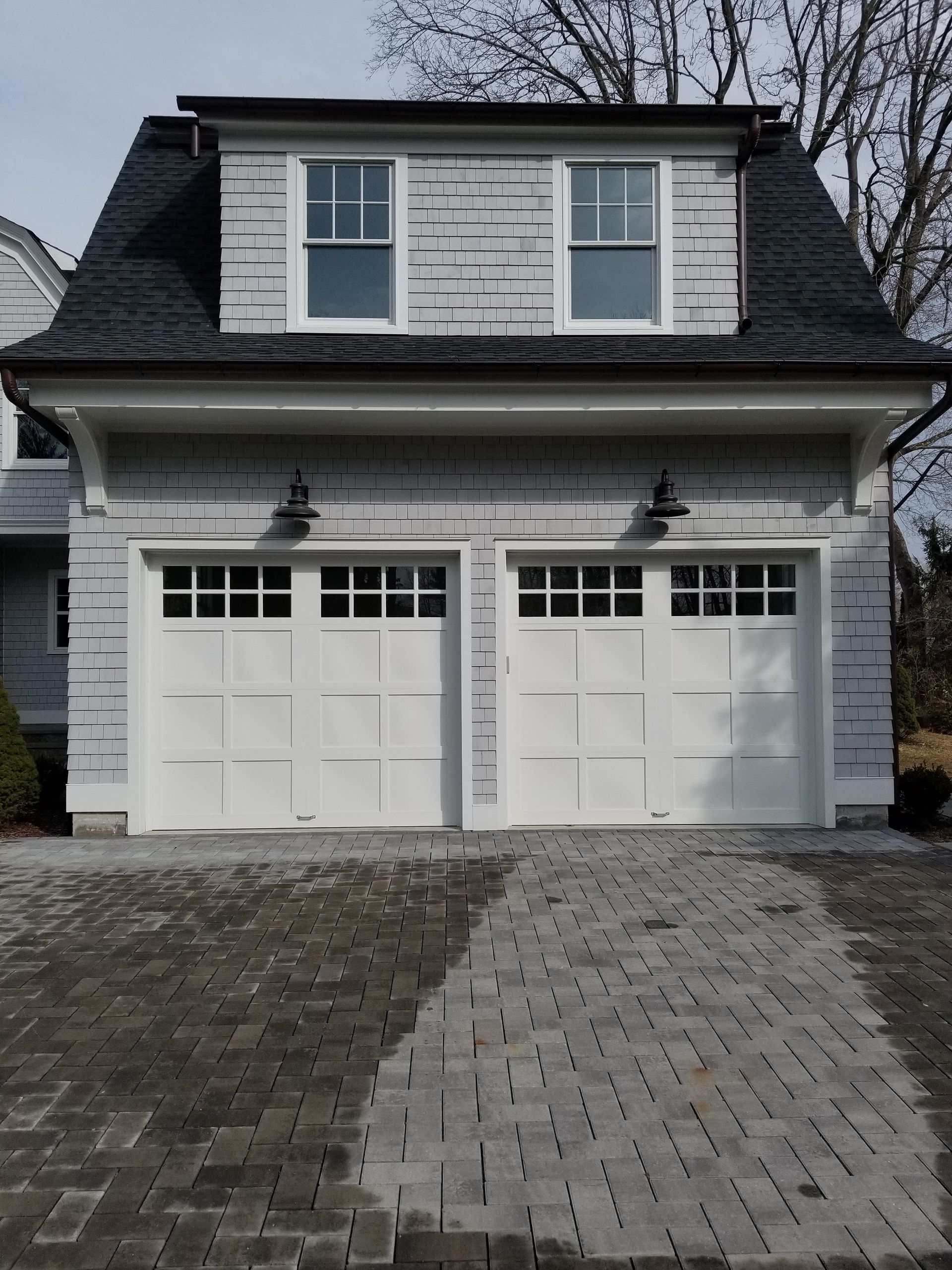 White two-car garage with dormer windows and a brick driveway under a cloudy sky