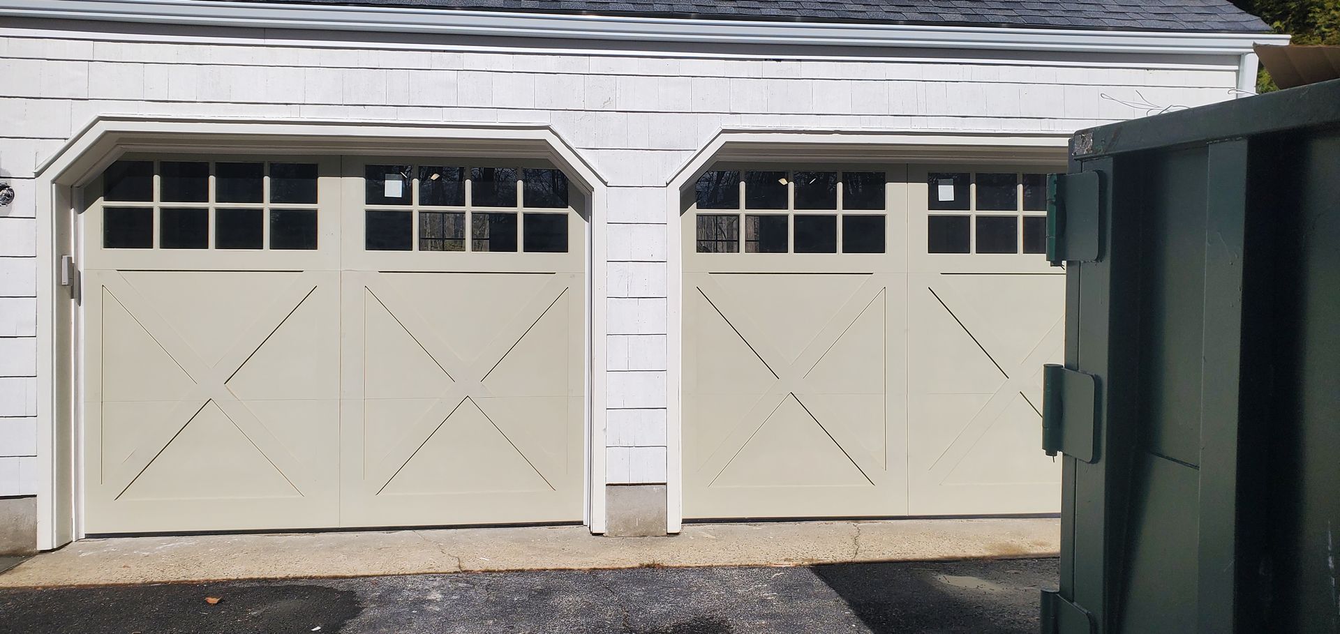 Two beige garage doors on a white house with a dark green dumpster at the right edge