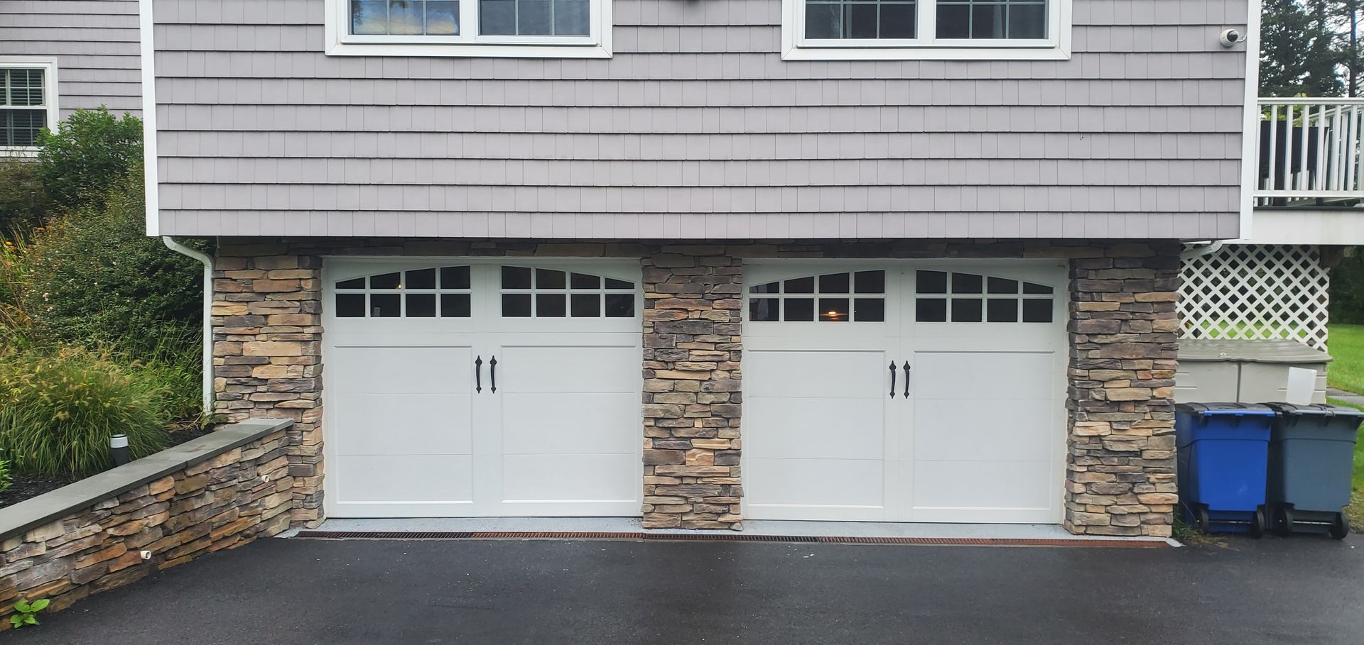 Two white garage doors beneath a gray house with stone trim and blue recycling bins