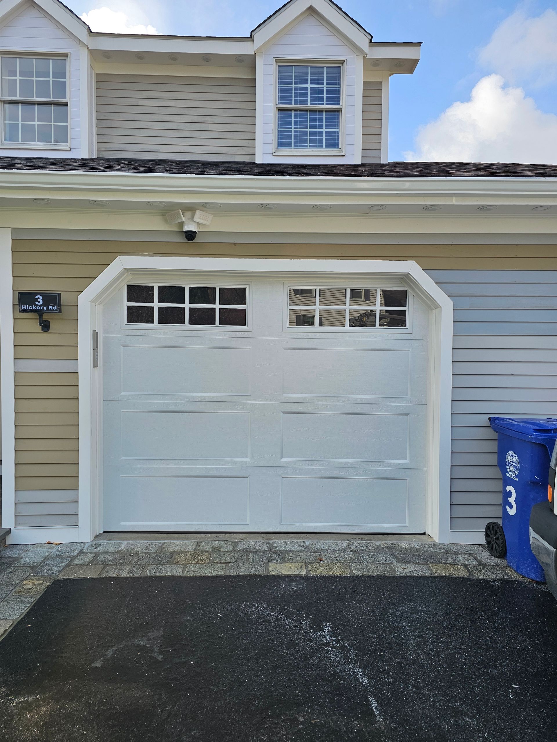 White garage door on a house with light gray siding and a blue recycling bin beside it.