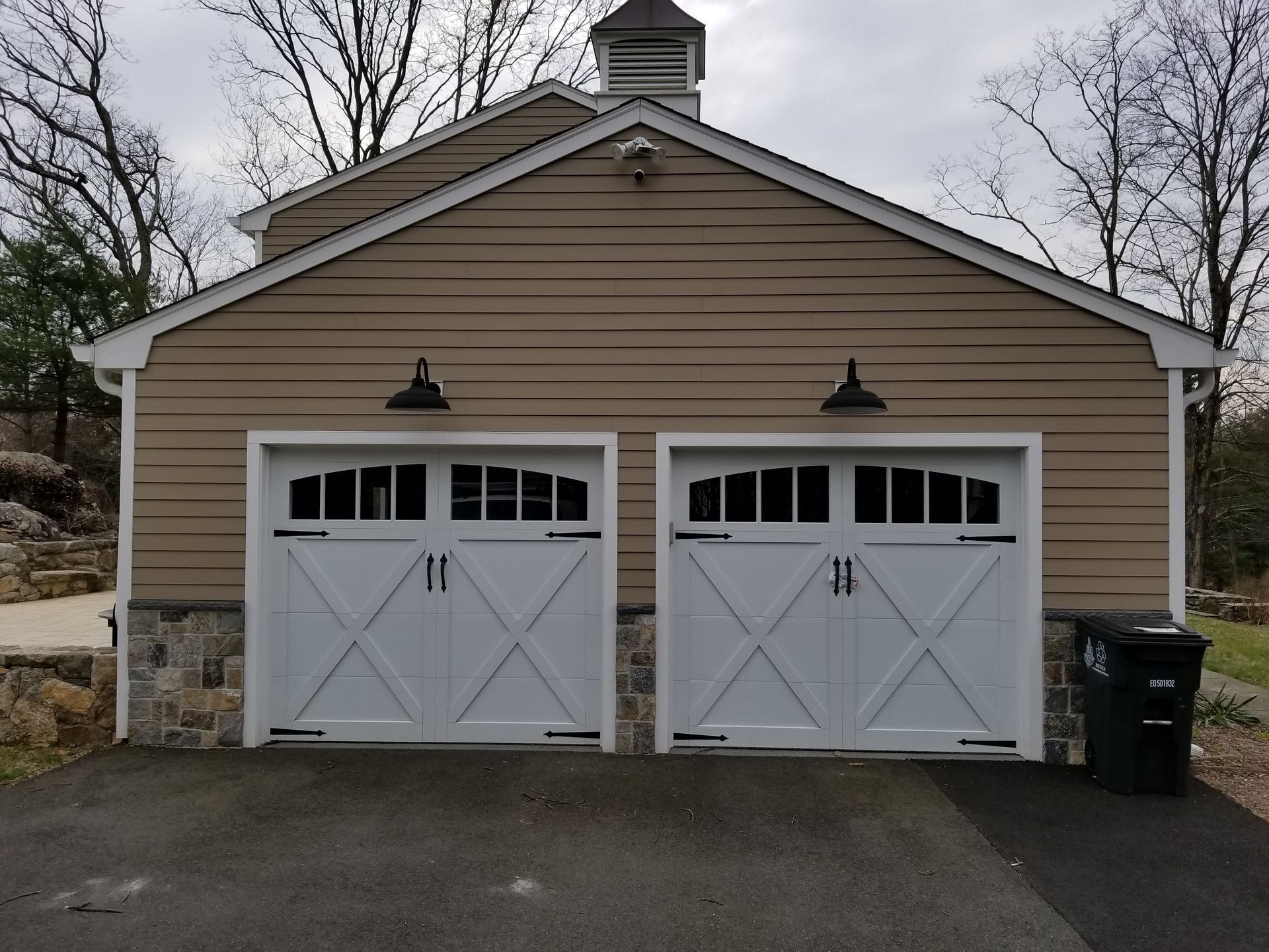 Two-car tan garage with white doors and black lantern lights under a gray sky