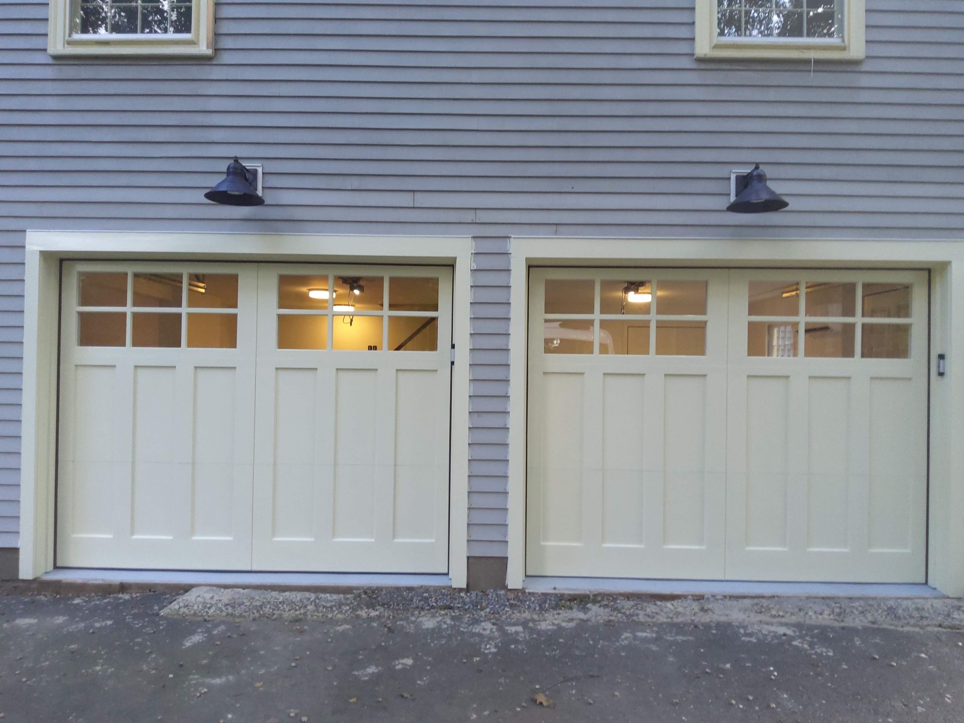 Two white garage doors on a gray-sided house, with exterior lights above each door.