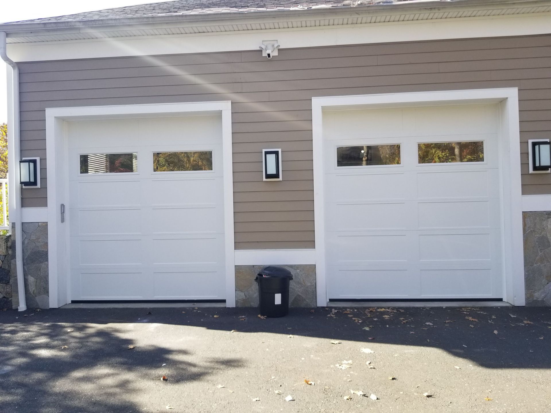 Two white garage doors on a tan house with black wall lights and a trash bin in front